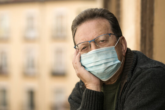 mature man in balcony home lockdown during covid19 outbreak - senior male on his 70s in face mask worried and tired looking to the street thoughtful and depressed in quarantine