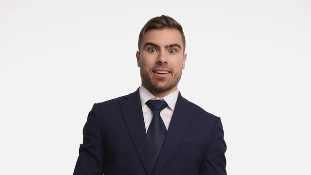 surprised attractive man in navy blue suit opening mouth and making thumbs up gesture, smiling and standing isolated on white background in studio