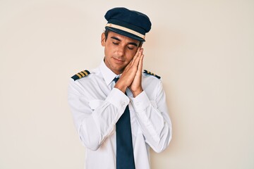 Young hispanic man wearing airplane pilot uniform sleeping tired dreaming and posing with hands together while smiling with closed eyes.