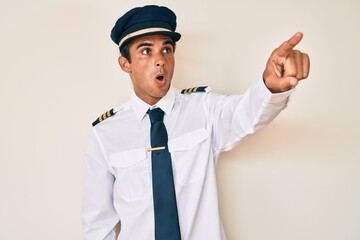 Young hispanic man wearing airplane pilot uniform pointing with finger surprised ahead, open mouth amazed expression, something on the front