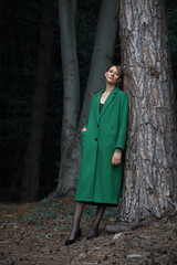 Fashionable young woman in green coat posing in a forest near the tree