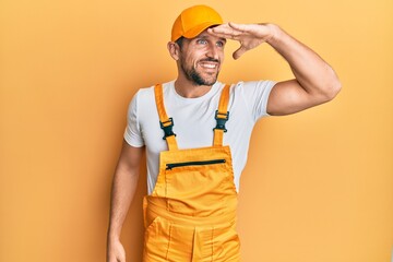 Young handsome man wearing handyman uniform over yellow background very happy and smiling looking far away with hand over head. searching concept.