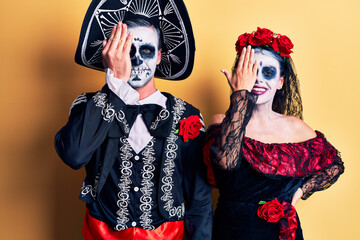 Young couple wearing mexican day of the dead costume over yellow covering one eye with hand, confident smile on face and surprise emotion.