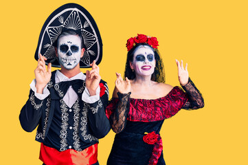 Young couple wearing mexican day of the dead costume over background gesturing finger crossed smiling with hope and eyes closed. luck and superstitious concept.