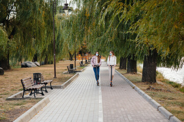 Two young beautiful girls in masks ride electric scooters in the Park on a warm autumn day. Walk in the Park.