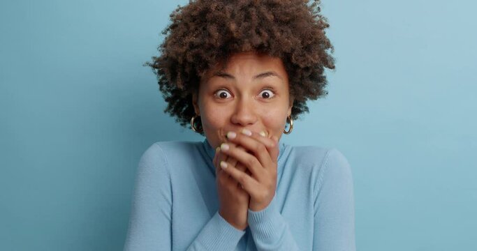 Pleased cheerful mysterious woman covers mouth and smiles gladfully looks surprisingly and hesitates shrugs shoulders wears long sleeved jumper isolated over blue background. Human reaction concept