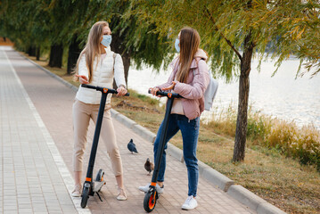 Naklejka na ściany i meble Two young beautiful girls in masks ride electric scooters in the Park on a warm autumn day. Walk in the Park.