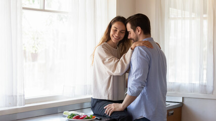 Cuddling for breakfast. Romantic millennial couple husband and wife hugging at kitchen at morning, young smiling woman girlfriend sitting on countertop hugging shoulders of affectionate man boyfriend