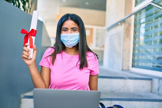 Young latin student girl wearing medical mask using laptop and holding diploma at university.