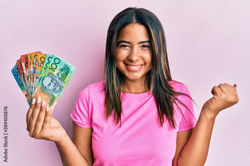 Poster Young latin girl holding australian dollars screaming proud, celebrating victory and success very excited with raised arm