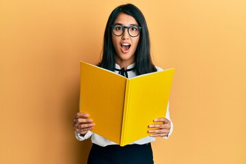 Beautiful asian young woman reading a book wearing glasses afraid and shocked with surprise and amazed expression, fear and excited face.