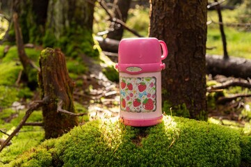 Pink baby thermos for drinks. Thermos in the forest on moss.