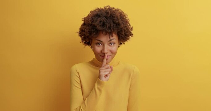 Happy mysterious woman with toothy smile puts index finger on lips has tricky gaze at camera tells secret only for you dressed in casual turtleneck isolated over yellow background spreads rumors