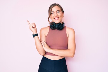 Beautiful young caucasian woman wearing gym clothes and using headphones smiling happy pointing with hand and finger to the side