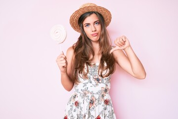 Beautiful young caucasian woman wearing summer hat and lollipop with angry face, negative sign...