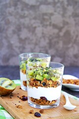 Homemade baked oat granola with nuts, dried cranberries and honey and unsweetened natural yogurt and kiwi in a glass glass on a dark concrete background. .