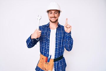 Young handsome man weaing handyman uniform holding wrench smiling with an idea or question pointing...