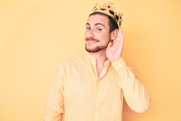 Young handsome man wearing king crown smiling with hand over ear listening and hearing to rumor or gossip. deafness concept.