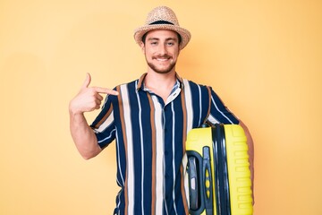Young handsome man wearing summer hat holding cabin bag pointing finger to one self smiling happy and proud