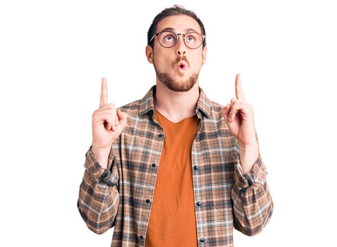 Young handsome caucasian man wearing casual clothes and glasses amazed and surprised looking up and pointing with fingers and raised arms.