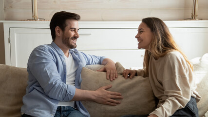 Resting at home together. Pleasant young millennial family couple, brother and sister, diverse friends sitting on cozy sofa at living room relaxing together talking joking having cheerful conversation