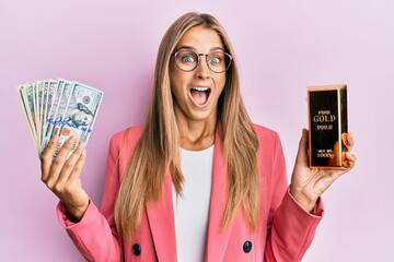 Young blonde woman wearing business style holding gold ingot and dollars celebrating crazy and...