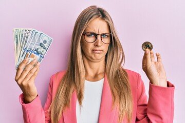 Young blonde woman wearing business style holding dollars and bitcoin skeptic and nervous, frowning upset because of problem. negative person.