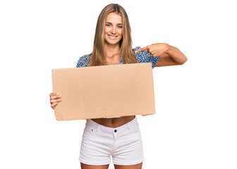 Young blonde woman holding cardboard empty banner pointing finger to one self smiling happy and proud