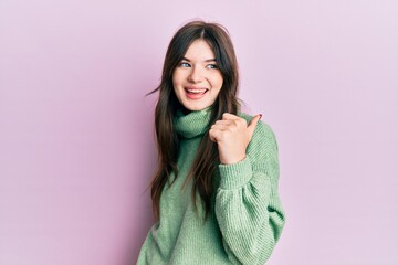 Young beautiful caucasian girl wearing wool sweater pointing thumb up to the side smiling happy with open mouth
