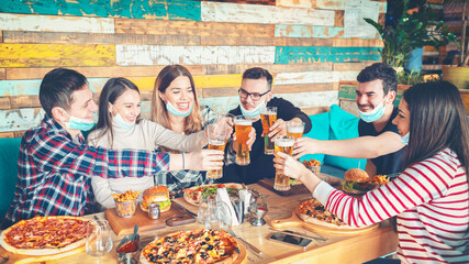 Happy young friends with protective face masks eating and drinking beer at restaurant
