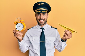 Handsome hispanic pilot man holding paper plane and alarm clock smiling with a happy and cool smile on face. showing teeth. - obrazy, fototapety, plakaty