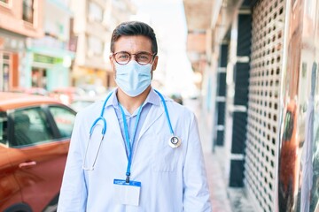 Young handsome hispanic doctor wearing uniform and coronavirus protection medical mask standing at town street.