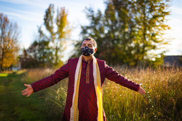 Mid aged person wearing traditional Indian clothes and face mask spreading hands for conveying embracing life in a meadow during sunset.