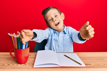 Adorable caucasian kid writing book sitting on the table looking at the camera smiling with open arms for hug. cheerful expression embracing happiness.
