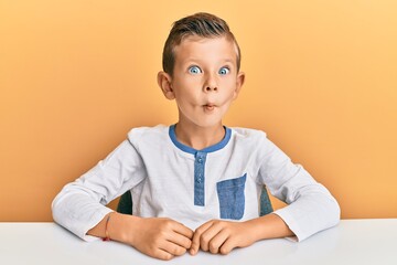 Adorable caucasian kid wearing casual clothes sitting on the table making fish face with lips, crazy and comical gesture. funny expression.