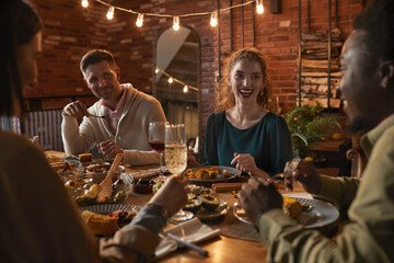 Group of cheerful adult people sitting at dinner table while enjoying party with outdoor lighting