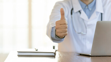 Doctor recommends. Close up of male physician therapist in lab coat sitting at desk in office by laptop showing thumb up gesture to camera advising proposing best clinic hospital service, copy space