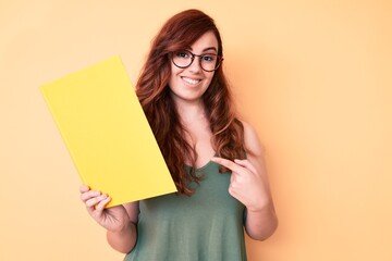 Naklejka premium Young beautiful woman wearing glasses holding book smiling happy pointing with hand and finger