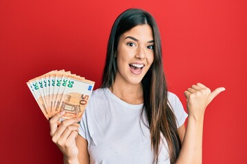 Young brunette woman holding 50 euro banknotes pointing thumb up to the side smiling happy with open mouth