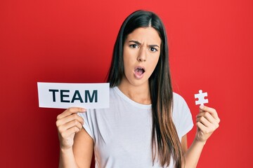 Young brunette woman holding team paper and piece of puzzle as teamwork in shock face, looking skeptical and sarcastic, surprised with open mouth