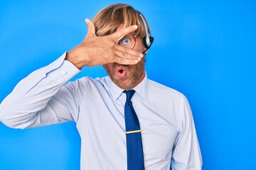 Young blond man wearing call center agent headset peeking in shock covering face and eyes with...