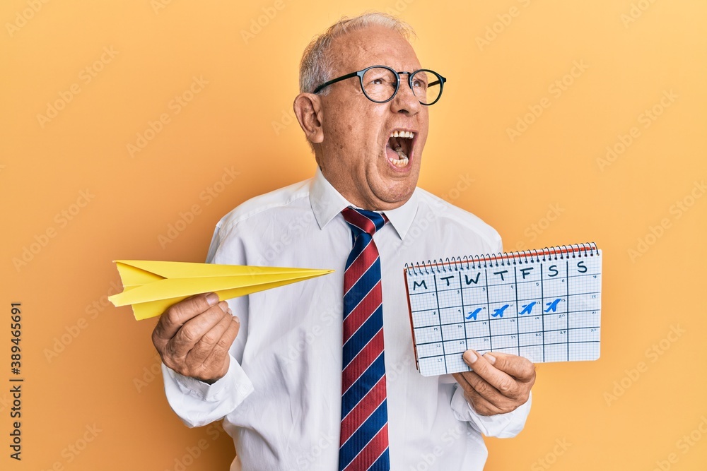 Sticker Senior caucasian man going on business trip holding paper airplane angry and mad screaming frustrated and furious, shouting with anger. rage and aggressive concept.