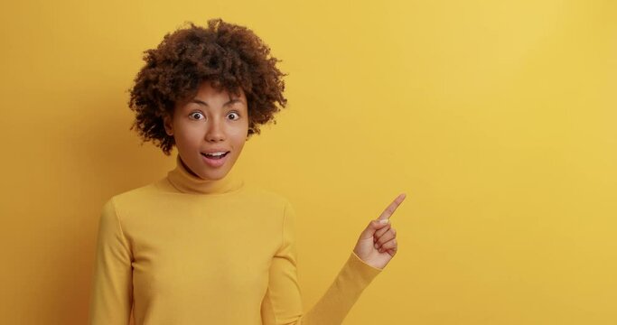 Video of pretty greatly surprised woman with Afro hair points right looks at something awesome with overjoyed expression says its unbelievable dressed in casual clothes shows blank space for promo - Powered by Adobe