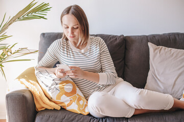 Young woman sitting on couch and texting talking at social media on smartphone. Middle age woman on sofa typing messaging on a mobile phone. Chat talk with friends online on Internet.