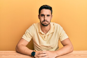 Young hispanic man wearing casual clothes sitting on the table puffing cheeks with funny face. mouth inflated with air, crazy expression.