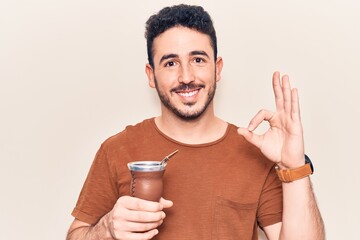 Young hispanic man drinking mate infusion doing ok sign with fingers, smiling friendly gesturing...