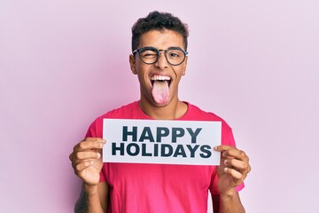 Young handsome african american man holding happy holidays message sticking tongue out happy with funny expression.