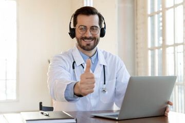 Perfect quality service. Portrait of happy doctor sitting at laptop in earphones showing thumb up...
