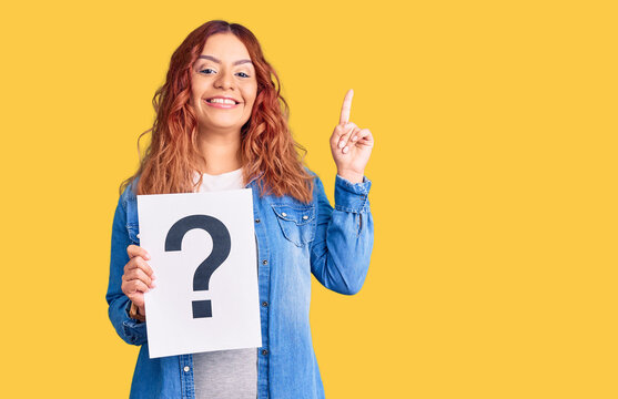 Young latin woman holding question mark surprised with an idea or question pointing finger with happy face, number one