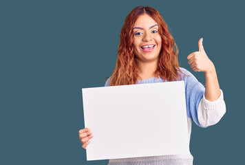 Fototapeta premium Young latin woman holding blank empty banner smiling happy and positive, thumb up doing excellent and approval sign